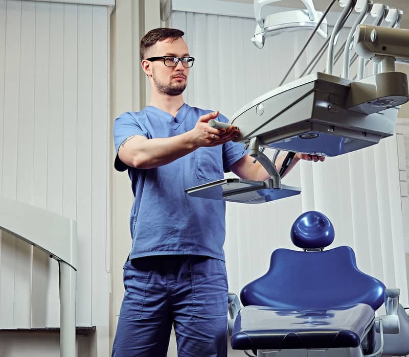 Male dentist in a room with medical equipment on background. רופא שיניים שיקום הפה